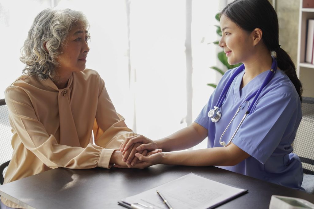 Nurse on right in scrubs holding the hands of an elder lady in a semi formal tan outfit.