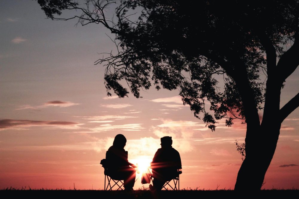 Two people sitting on chairs a sunset in the background with a tree on their right.